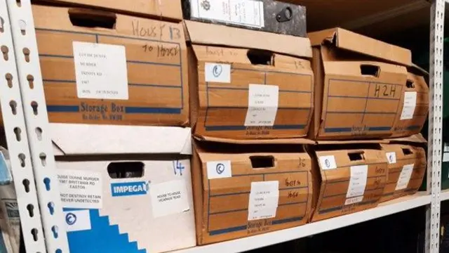 Several cardboard boxes and a large file stored on a shelving unit. The boxes are all labelled and many have writing on them.