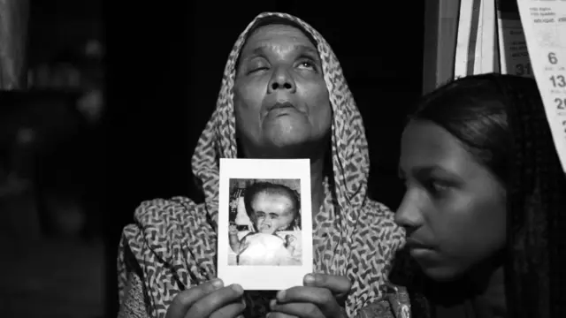 জায়নাবাকে ধরে আছেন তার মা জামিলা। তার হাইড্রোসেফালাস হয়েছিল। এক বছর বয়স পূর্ণ হওয়ার আগেই ২০০১ সালে মারা যায় শিশু জায়নাবা। মি. মধুরাজ যখন ২০১০ সালে তাদের বাড়িতে যান, তখন মেয়ের এই ছবিটি বার করেছিলেন মিজ. জামিলা। যত্ন করে একটা খামের মধ্যে ছবিটা রেখে দিয়েছিলেন তিনি।