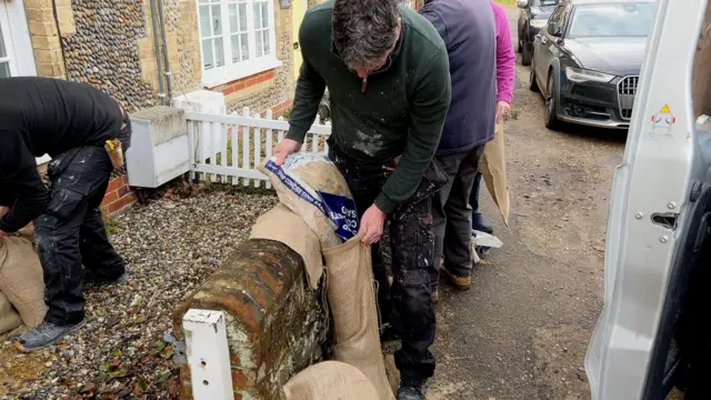 Un hombre con un jersey verde y pantalones negros pintados vierte arena en una bolsa fuera de una casa. Al fondo, otras personas hacen lo mismo.