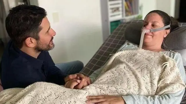 Paola Roldán, sentada na cama, com um respirador no nariz, coberta com um cobertor e segurando a mão do marido. 