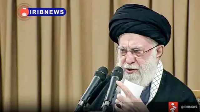 İran'ın dini lideri Ali Hamaney iç mekanda mikrofonlara doğru konuşma yapıyor. Yalnızca göğüsten yukarısı görülüyor. Üzerinde siyah cübbe ve sarık var.