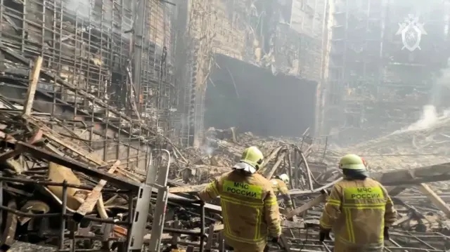 Two emergency workers in yellow jacks and hats in front of the burnt out remains of the Crocus City Hall concert venue, 23 March 23 2024
