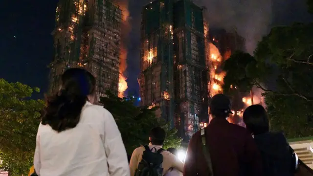 Люди наблюдают за густым дымом и пламенем, поднимающимся во время пожара в жилом комплексе Wang Fuk Court в Гонконге 26 ноября 2025 года.