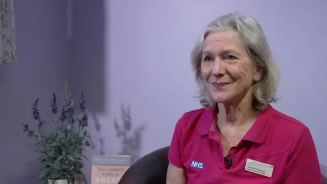 Una mujer de pelo largo y canoso, con una camiseta rosa del NHS, sonríe. Lleva una insignia del NHS en la parte superior y un fondo morado con una planta a su derecha.