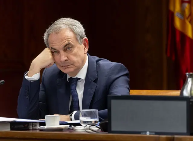 Zapatero, sentado en una sala institucional, apoyando la cabeza en la mano con micrófono, taza y vaso, con una bandera al fondo.