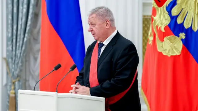 Мужчина с алой наградной лентой через плечо на трибуне перед двумя микрофонами. На заднем плане российские флаги