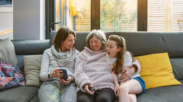 Três gerações de mulheres de uma mesma família sentadas no sofá, observando uma foto no celular