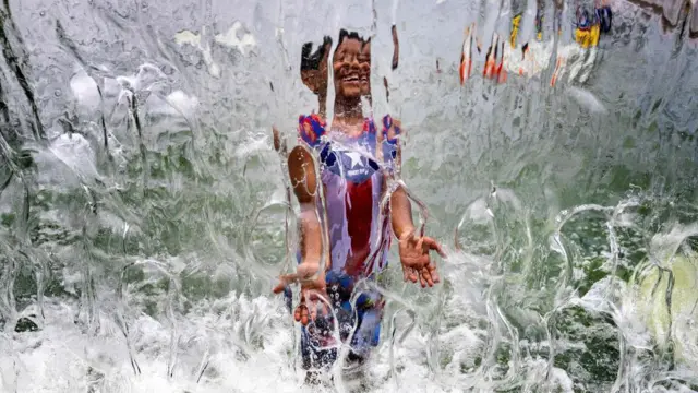 美國東岸的熱浪中，一名小男孩在噴泉下乘涼——水的反射使照片失真。
