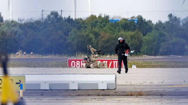 黒い長袖長ズボン姿の人物が、空港の駐機場を歩いている。右隣にベージュのロボットが見える