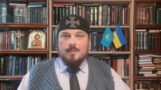 Воронцов сидит на фоне книжных полок, слева от него стоит икона, а справа флаги Казахстана и Украины. На голове у него черная шапочка с вышитым крестом