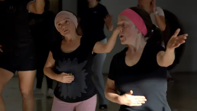 María Luisa ve María del Cisne bir dans dersinde - ikisi de bir ellerini karınlarının üzerine koymuş, diğer ellerini havada tutuyorlar. İkisi de siyah tişört ve pembe saç bandı takıyor.