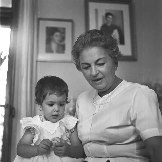 Cristina con una blusa blanca de manga corta ve hacia abajo mientras tiene a su lado a la pequeña nieta, que lleva un vestido blanco sin mangas y mira en la misma dirección que su abuela. Están en una habitación, en el fondo se pueden ver dos retratos de mujeres colgados en la pared, uno de ellos es de Frida Kahlo.