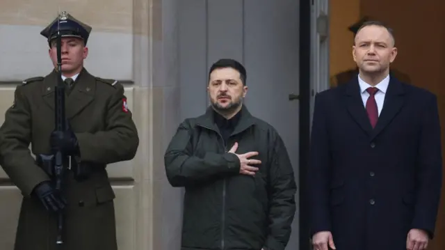 Володимир Зеленський у темно-зеленій куртці, він тримає руку на серці і Кароль Навроцький у темно-синьому пальто, вони слухають гімни країн перед початком зустрічі