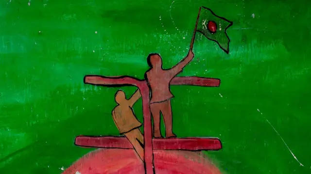 Um mural mostra o contorno de duas pessoas sobre uma estrutura, acenando uma bandeira, diante de um fundo com um nascer do sol nas cores da bandeira de Bangladesh