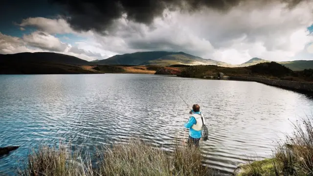 Hombre pescando frente a pequeño lago. Al fondo nubes sobre las montañas.
