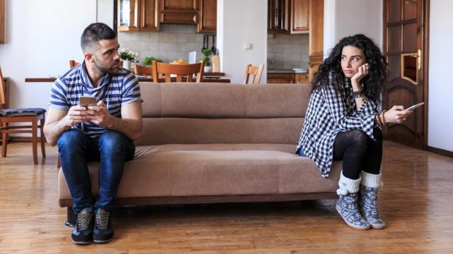 Um homem e uma mulher sentadosbrasileirao serieum sofá, cada umbrasileirao serieum canto, ambos mexendo no celular