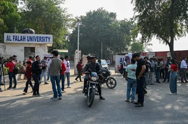 হরিয়ানার আল-ফালাহ বিশ্ববিদ্যালয়ের সামনে দাঁড়িয়ে আছে বেশকিছু মানুষ। দুটি মোটরসাইকেল যাচ্ছে সেখান দিয়ে 