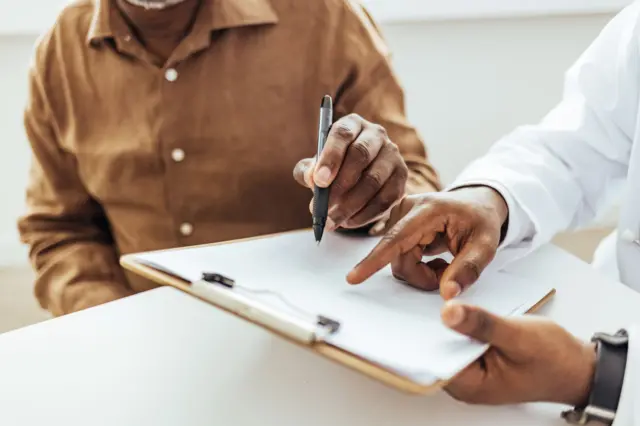 Un paciente se dispone a firmar un documento acompañado de otra persona que lo sujeta