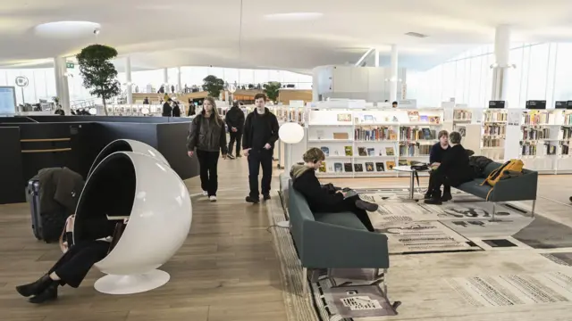 Interior de uma biblioteca moderna, com iluminação natural e grandes janelas. Há estantes baixas cheias de livros organizados, sofás e mesas onde pessoas leem, conversam ou usam dispositivos eletrônicos. À esquerda, uma pessoa está sentada dentro de uma cadeira em formato de cápsula branca, parcialmente fechada. No centro, duas pessoas caminham pelo espaço. O ambiente tem design contemporâneo, com piso de madeira, tapetes e plantas distribuídas pelo local.