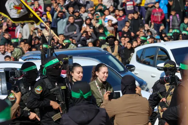 Hamas fighters, dressed in black with their faces covered and wearing green bandanas, escort two Israeli hostages as a crowd watches on