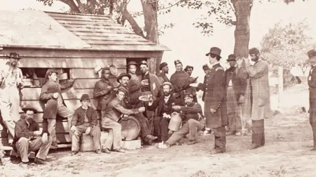 Un grupo de soldados de la guerra de secesión está bebiendo frente una casa de madera. 