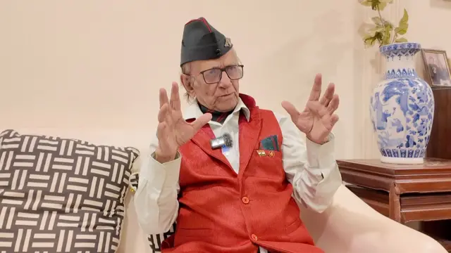 সাদা শার্ট, লাল জ্যাকেট ও মাথায় কালো টুপি পরে দুই হাত তুলে কথা বলছেন দীপঙ্কর ব্যানার্জী