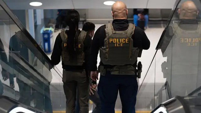 Two US Immigration and Customs Enforcement agents, one with a shaved head and wearing bullet-proof vests labelled "POLICE ICE", stand on a descending escalator.