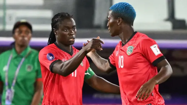 Tabitha Chawinga tend la main droite pour serrer celle de Temwa Chawinga, de profil et dos à l'objectif. Les deux joueuses portent le maillot rouge du Malawi à liserés noirs : Tabitha arbore un brassard de capitaine vert et le numéro 11 blanc, tandis que Temwa a les cheveux teints en bleu et le numéro 10. À l'arrière-plan, on aperçoit, flous, un entraîneur en maillot vert, un banc de touche et des tribunes clairsemées.