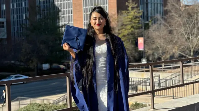 شکیلا محمدی با لباس فراغت دانشگاه، در حالی که کلاه فارغ‌التحصیلی را در دست گرفته است، با لبخند به کمره نگاه می‌کند. 