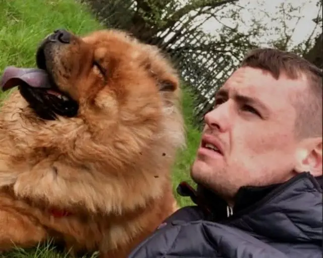 Чоловік, що дивиться вбік від камери, стоїть поруч із собакою породи чау-чау коричневого кольору на галявині. У собаки висунутий язик.