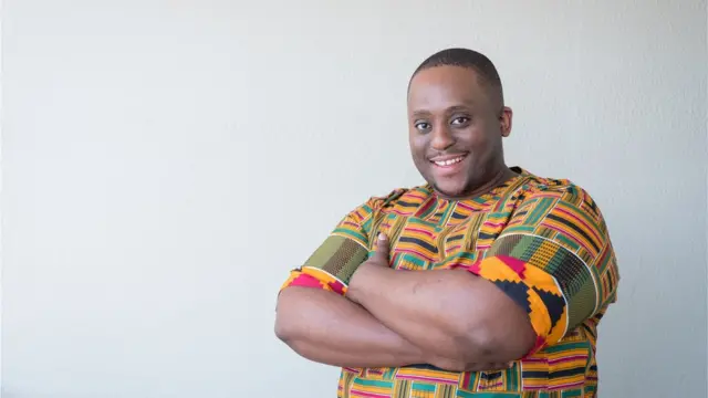 Portrait d'un homme souriant en surpoids portant des vêtements traditionnels. L'homme a les bras croisés contre le mur de sa maison. Il a un regard confiant sur son visage.