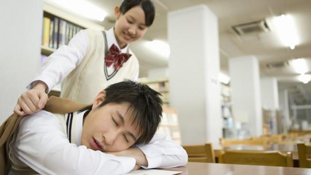 مراهق نائم - رأسه على الطاولة والكتب - في المكتبة؛ وفتاة عابرة تضع معطفا على كتفيه