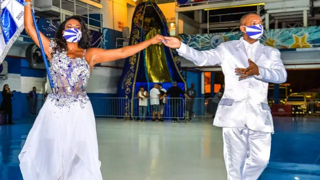 Um homem e uma mulher, usando máscaras, dançam em uma quadra vazia; ela segura bandeira da escola de samba Beija-Flor de nilópolis