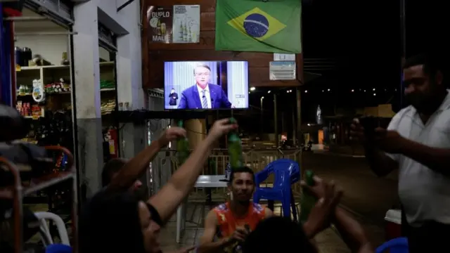 Bolsonaro na tela TVh2bet ajudabar, com pessoas conversando a frente