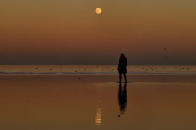 A woman looks at a full Moon while walking in the water at Seapoint beach in Dublin, Ireland, on 17 January 2022