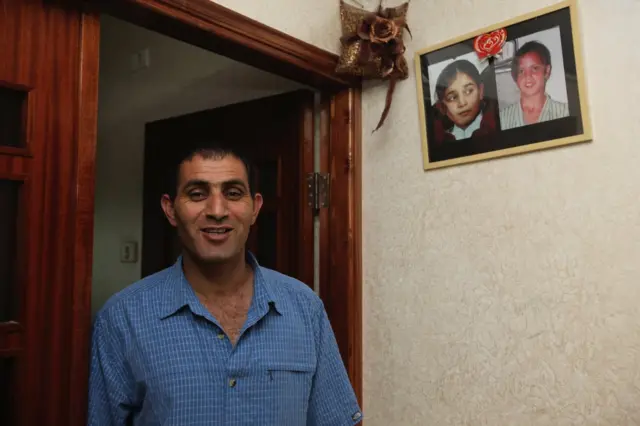 Bassam Aramin standing in a doorway with photos of his daughter and Ram's daughter hanging on his wall