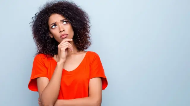 A young woman looks doubtful, she's got her arms crossed and is holding her chin with one hand. Pale blue background.