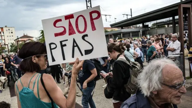 یوه ښځه د ګڼې، ګوڼې په منځ کې ولاړه ده او د کیمرې پر لور یې شا کړې ده. هغه کاغذ، چې هغې په لاس کې نیولی دی، پرې لیکل شوي "PFAS ودروئ." هغه تور ویښتان لري او نیلي رنګه جامې یې اغوستي دي.