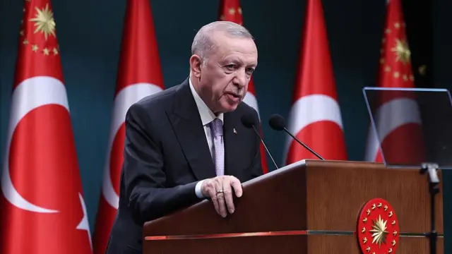 د ترکیې ولسمشر رجب طیب اردوغان، چې دریشي او نیک ټاي اغوستې، د ترکیې بېرغ مخې ته وینا کوي.