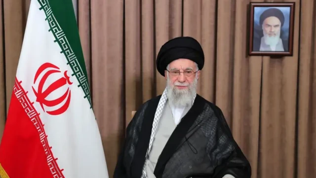 علی خامنه‌ای و پرچم جمهوری اسلامی در کنارش، تصویر روح الله خمینی در پشت سر او روی پرده قهوه‌ای رنگ نصب شده است