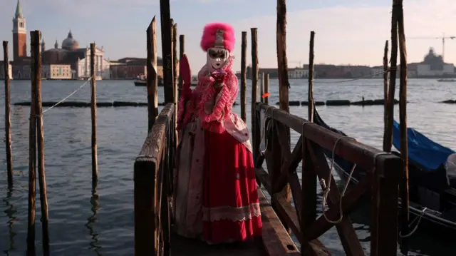 Devojka u dugoj haljini roze boje, lice joj prekriva bela maska, a na glavi ima šubaru koja je takođe roze boje