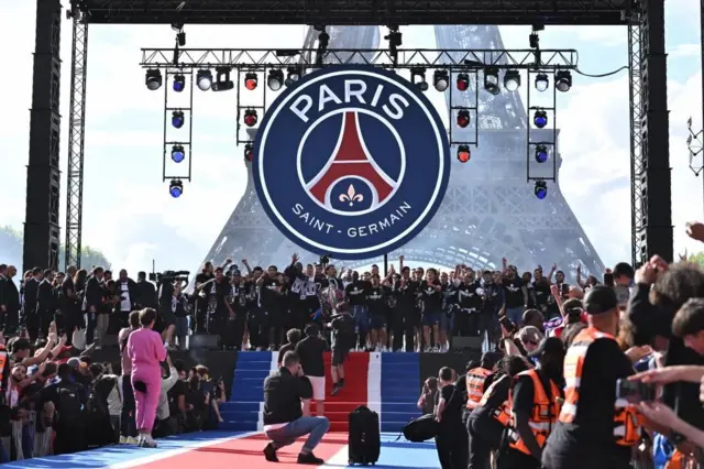 エッフェル塔の前に設けられた舞台に、「PARIS Saint-Germain」と書かれたチームのエンブレムが掲げられ、壇上につながる通路には同じ青赤白のじゅうたんがしかれている。檀上では選手たちが集まり喜び、手前の左右には大勢のファンが集まり、オレンジ色のベストを着た警備員たちが前に出ないよう押しとどめている。