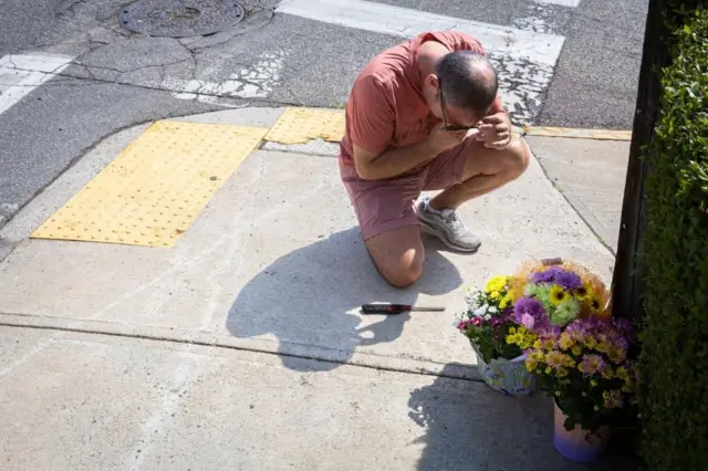交差点の角の歩道に、複数の花束が置かれ、その前にピンク色のTシャツとショーツ姿の人がひざまずき、口元を手で押さえている