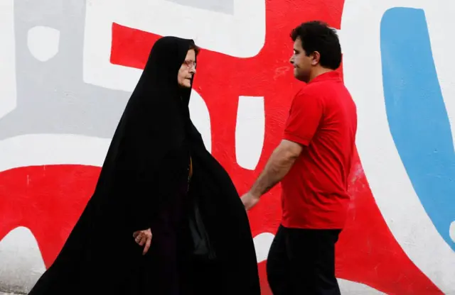 مردی با تی‌شرت قرمز و زنی با چادر مشکی در جهت مخالف در پیاده‌رو حرکت می‌کنند