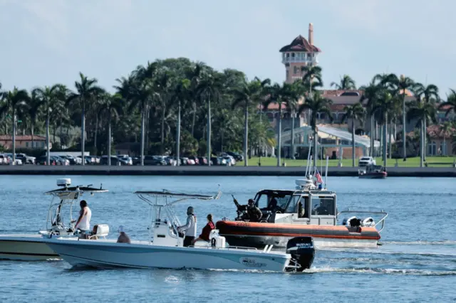 Водойма з кількома моторними човнами на передньому плані. Один човен білий, з відкритою палубою, на ньому кілька людей. Поруч – інший човен сірого та помаранчевого кольору, виглядає як патрульний або охоронний, з обладнанням і людьми на борту. На березі – рівний ряд високих пальм, а за ними – велика будівля з червоним дахом і баштою, що нагадує розкішний маєток або клуб. Погода сонячна, вода спокійна, небо ясне.