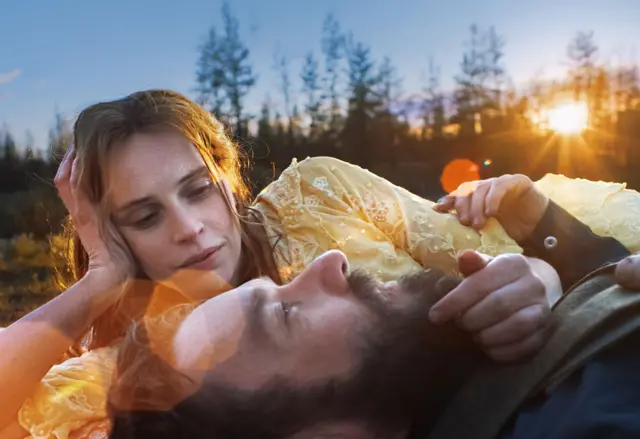 A imagem mostra um homem e uma mulher deitados na grama ao ar livre, iluminados pela luz dourada do pôr do sol. A mulher, com cabelos claros e vestido amarelo delicado, apoia a cabeça na mão enquanto olha com ternura para o homem, que está deitado de costas, com a cabeça apoiada próxima a ela. Ele tem barba e cabelo castanho, e segura levemente o braço dela. Ao fundo, há árvores em silhueta e o sol baixo no horizonte cria reflexos alaranjados na lente, conferindo à cena uma atmosfera íntima, tranquila e contemplativa.