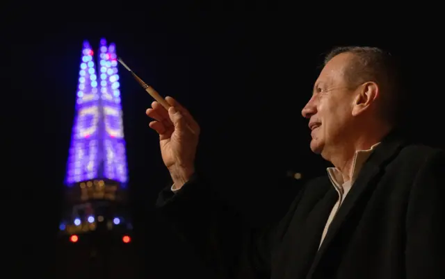 Una imagen del pintor Gustavo Zuluaga con un pincel en la mano apuntando a la torre The Shard, que se ve iluminada en el fondo de la foto.