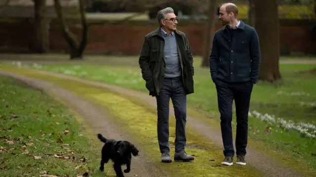 Юджин Леви и принц Уильям в осенней одежде прогуливаются по парку, рядом с ними бежит небольшая черная собака.