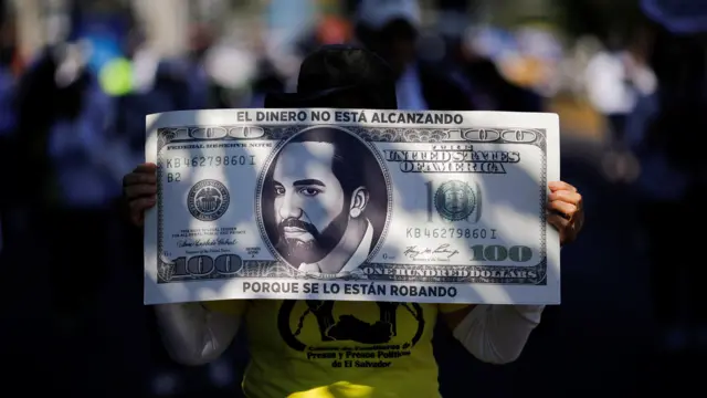 Una manifestante sostiene un cartel que representa un billete de 100 dólares con el rostro del presidente Nayib Bukele impreso y el mensaje 