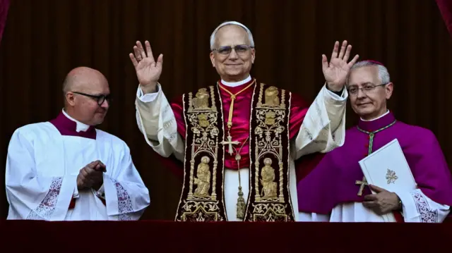 Pope Leo XIV stand for balcony bifor di crowd for St Peter's Square, e hold im hands up alongside two Vatican officials